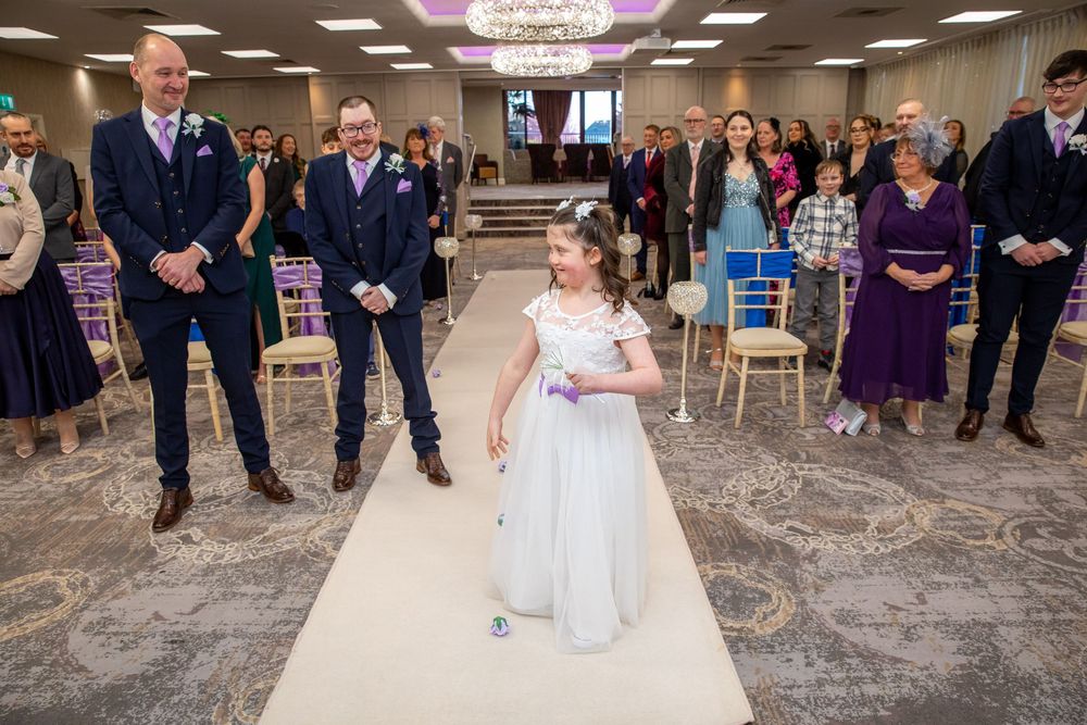 Flower girl in white dress walks down wedding aisle as guests watch during indoor ceremony.