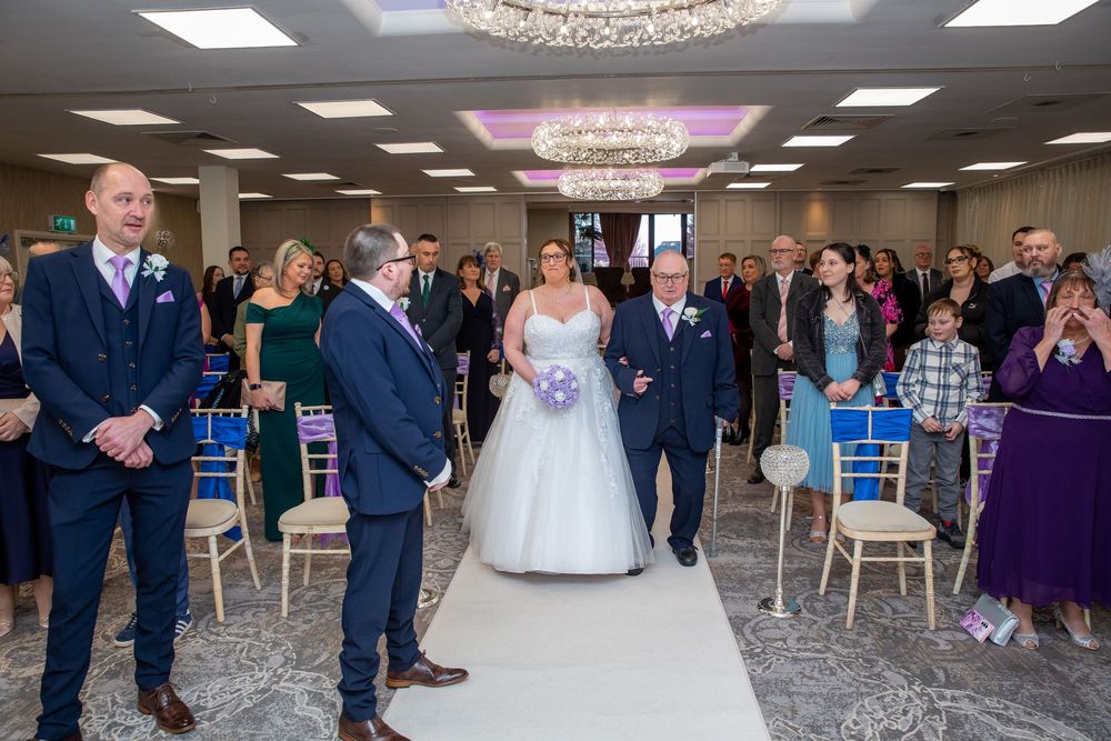 Bride walks down the aisle with her father at an elegant wedding ceremony with blue chairs and chandelier lighting.