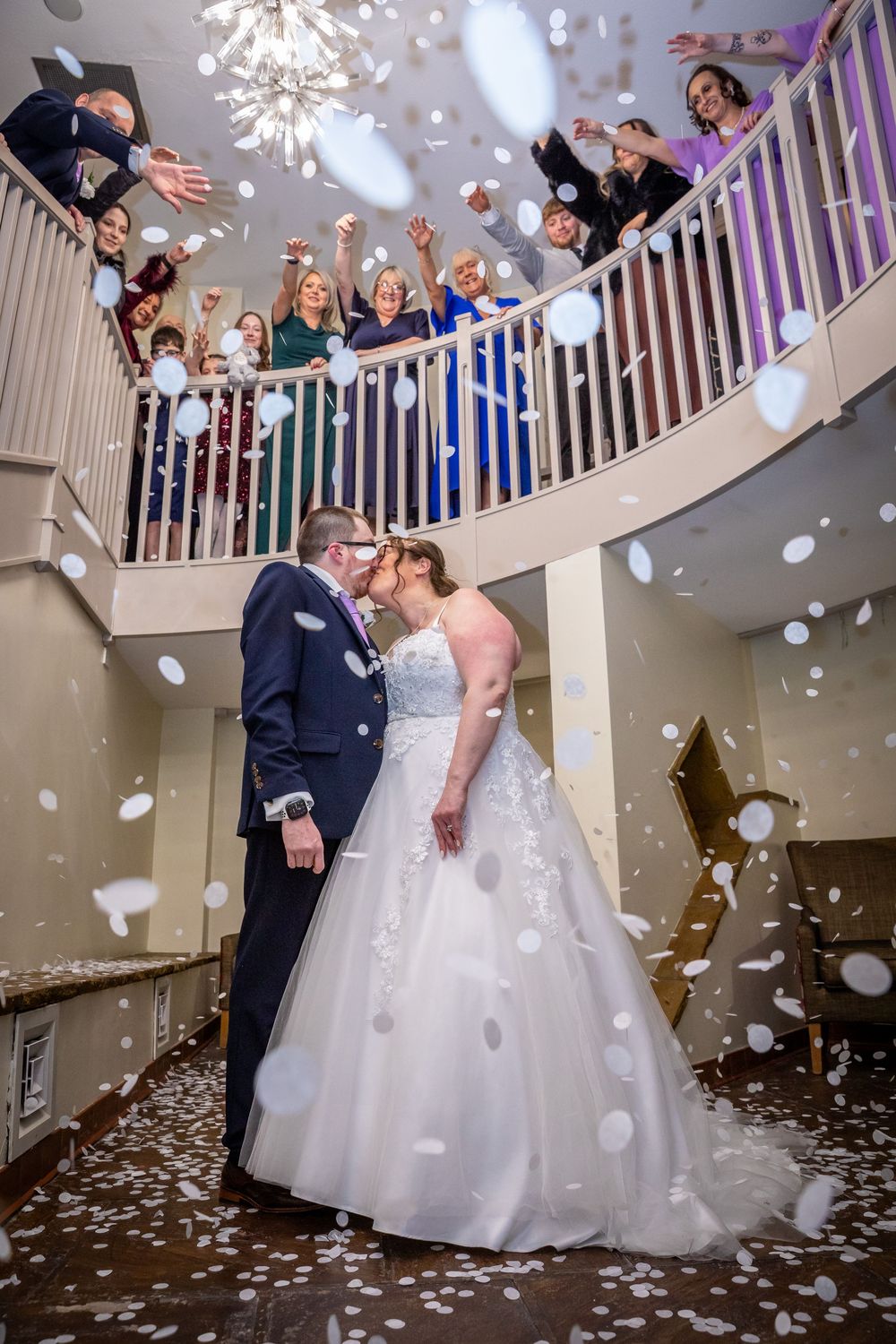 Bride and groom share first kiss under falling white confetti while wedding guests cheer from a curved staircase above.