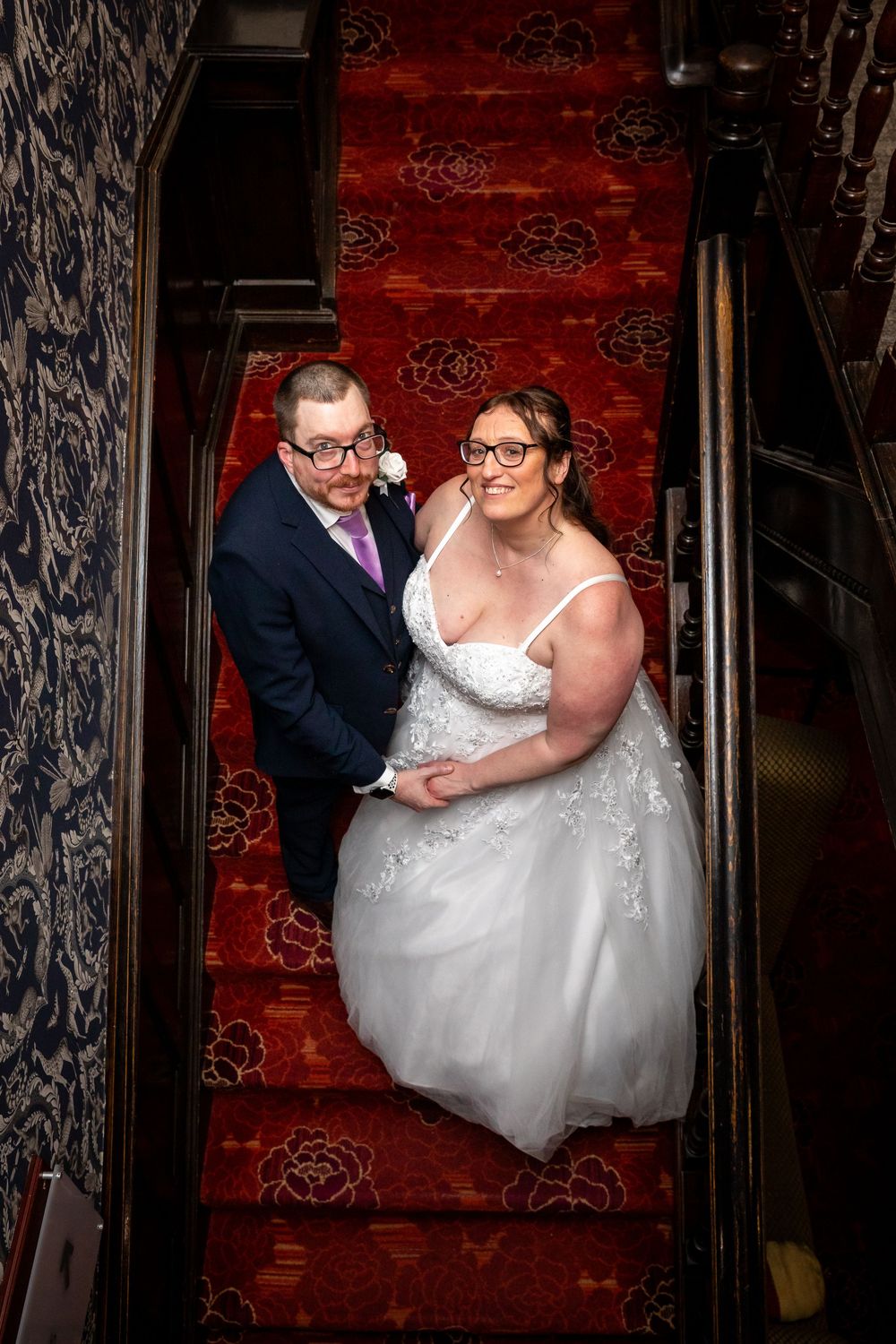 Bride and groom posing on a red carpeted staircase, she in a white gown and he in a navy suit with purple tie.