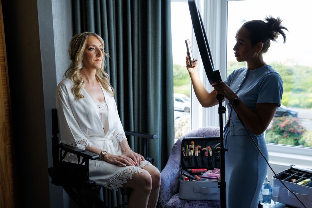 Bride in white robe gets hair styled by makeup artist near window in hotel room before wedding.