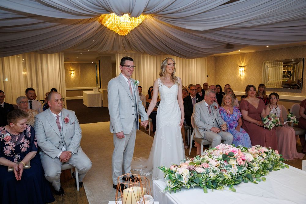 Bride and groom holding hands at indoor wedding ceremony with seated guests in elegant decorated venue.