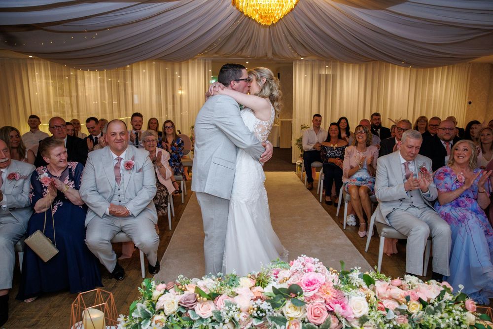 Bride and groom share first kiss at wedding ceremony with guests applauding in decorated venue with floral arrangements.
