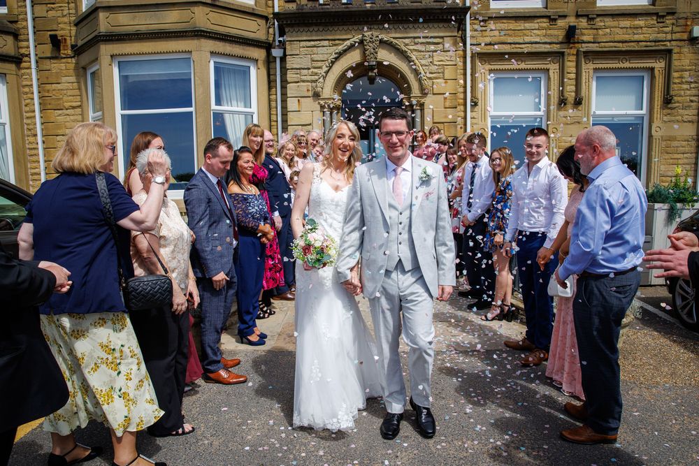 Bride and groom walk through confetti shower outside venue, surrounded by smiling wedding guests on a sunny day.