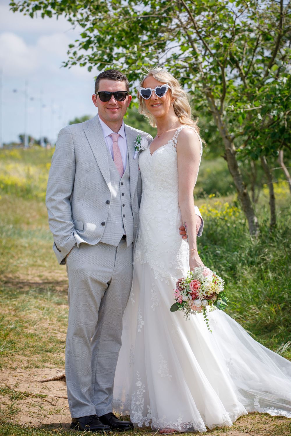 Bride and groom posing outdoors in summer, wearing sunglasses, grey suit and lace wedding dress, holding floral bouquet.