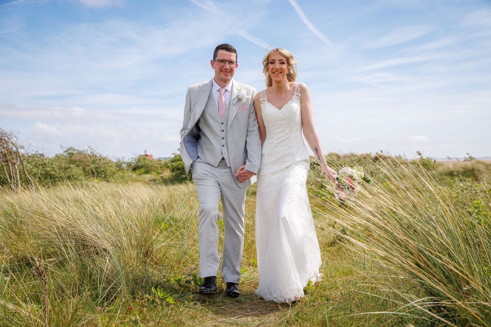 Bride and groom holding hands walking through tall grass on a sunny day at an outdoor wedding venue.