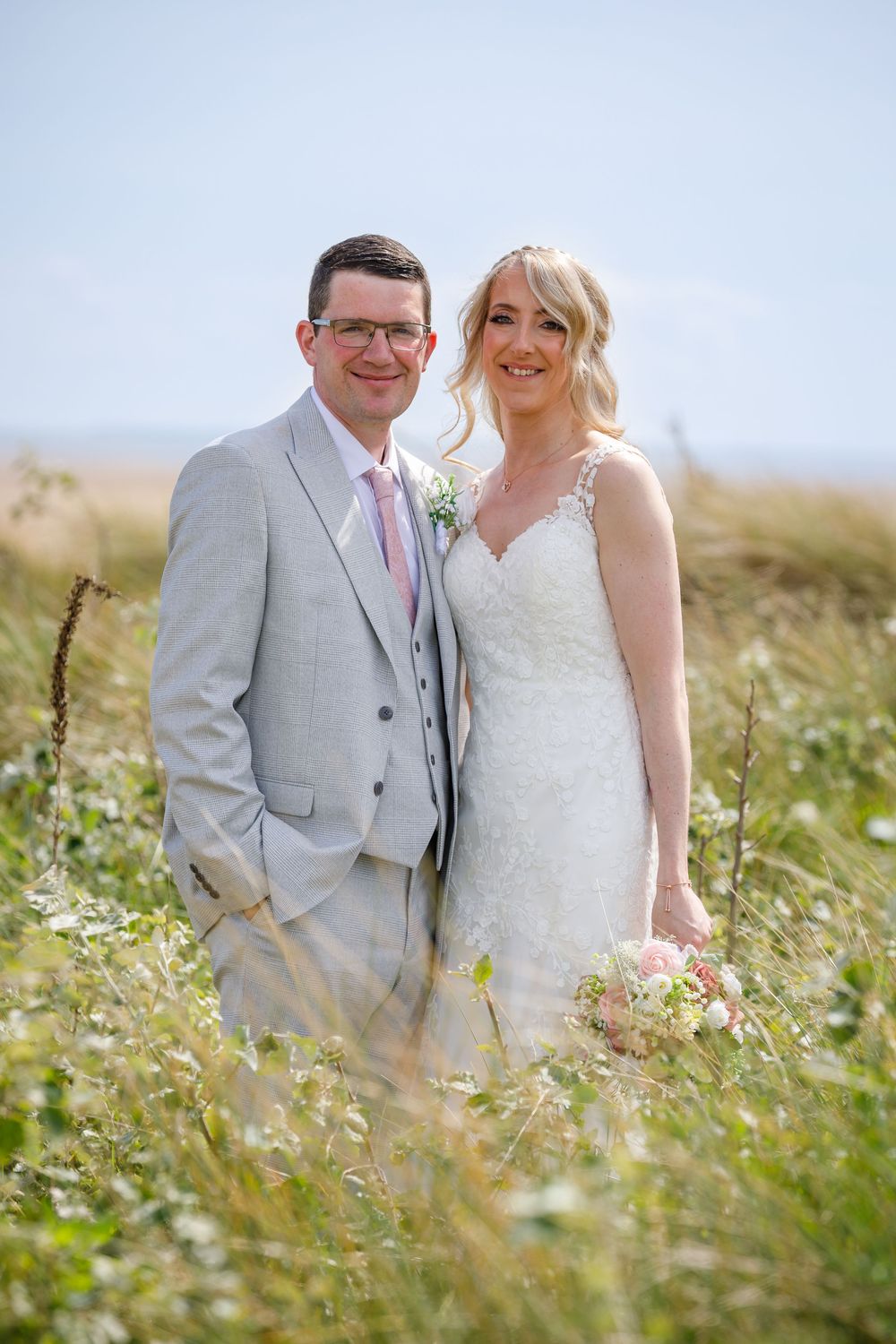 Bride and groom smiling in a wildflower meadow, bride in lace gown holding pink bouquet, groom in grey suit with pink tie.