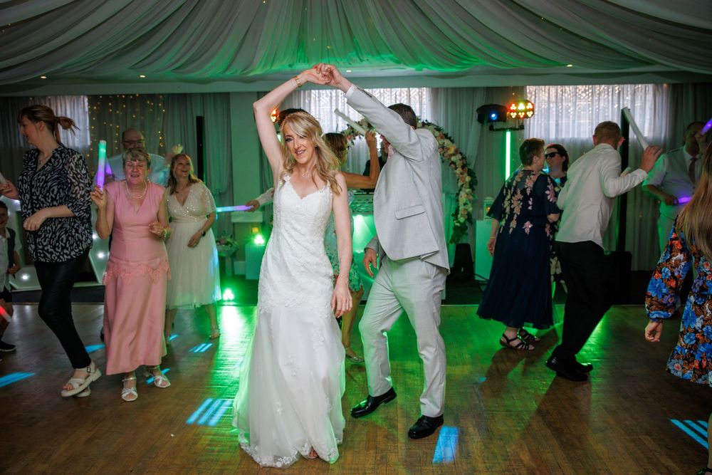 Bride and groom dancing together at wedding reception with guests on dance floor under green lights.