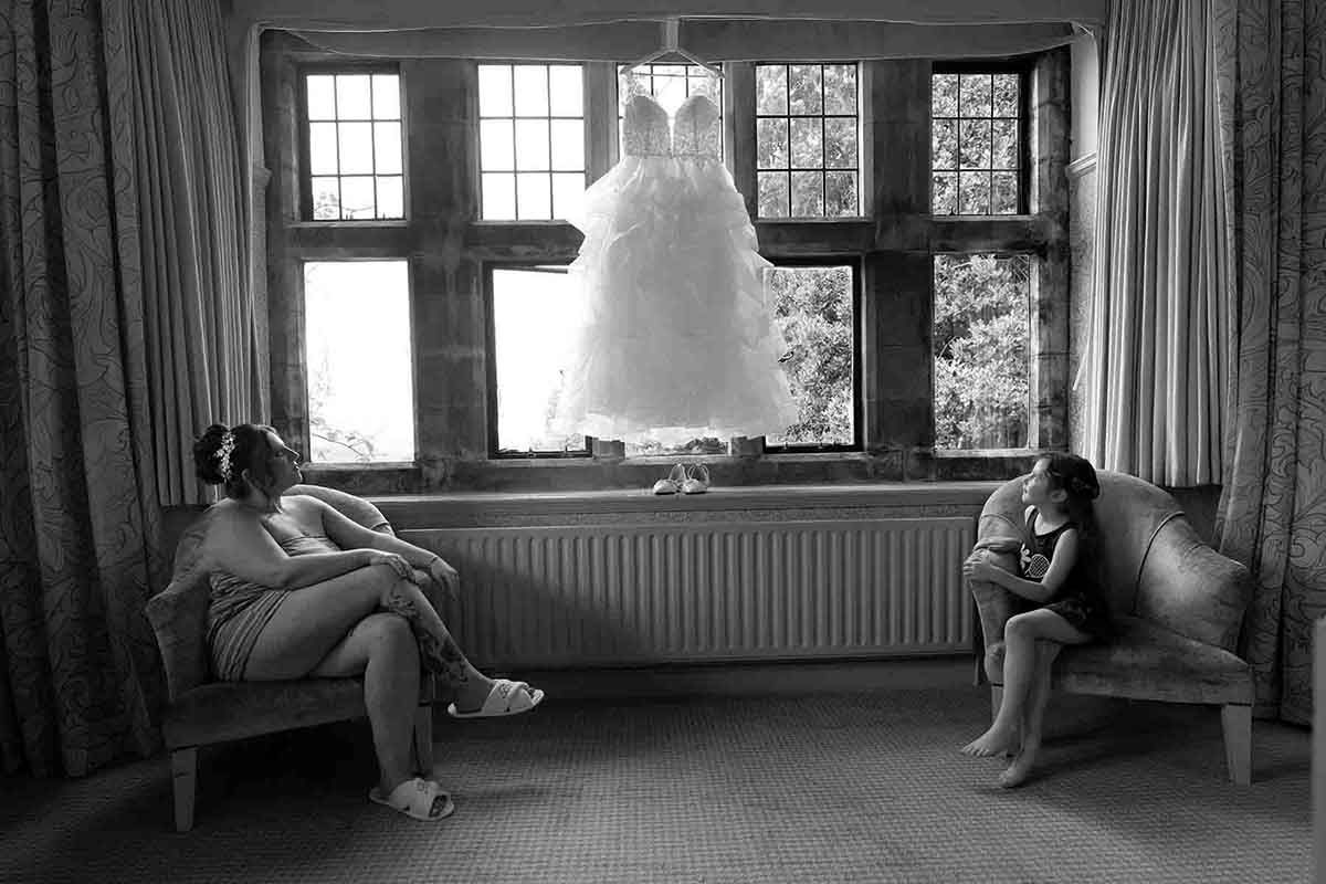 Black and white photo of two women relaxing on sofas with a wedding dress hanging in a sunlit window.
