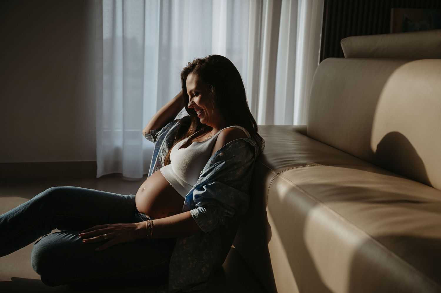 Pregnant woman in jeans and plaid shirt sitting on floor leaning against sofa, cradling baby bump in natural light.