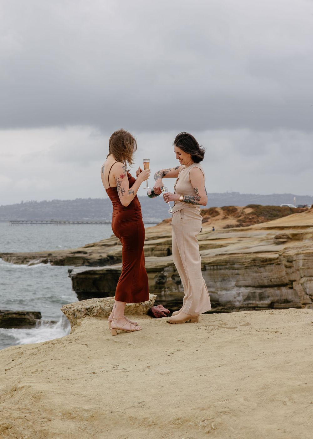 Two women standing on rocky coastal cliffs holding champagne glasses, one in red dress, one in beige pants.