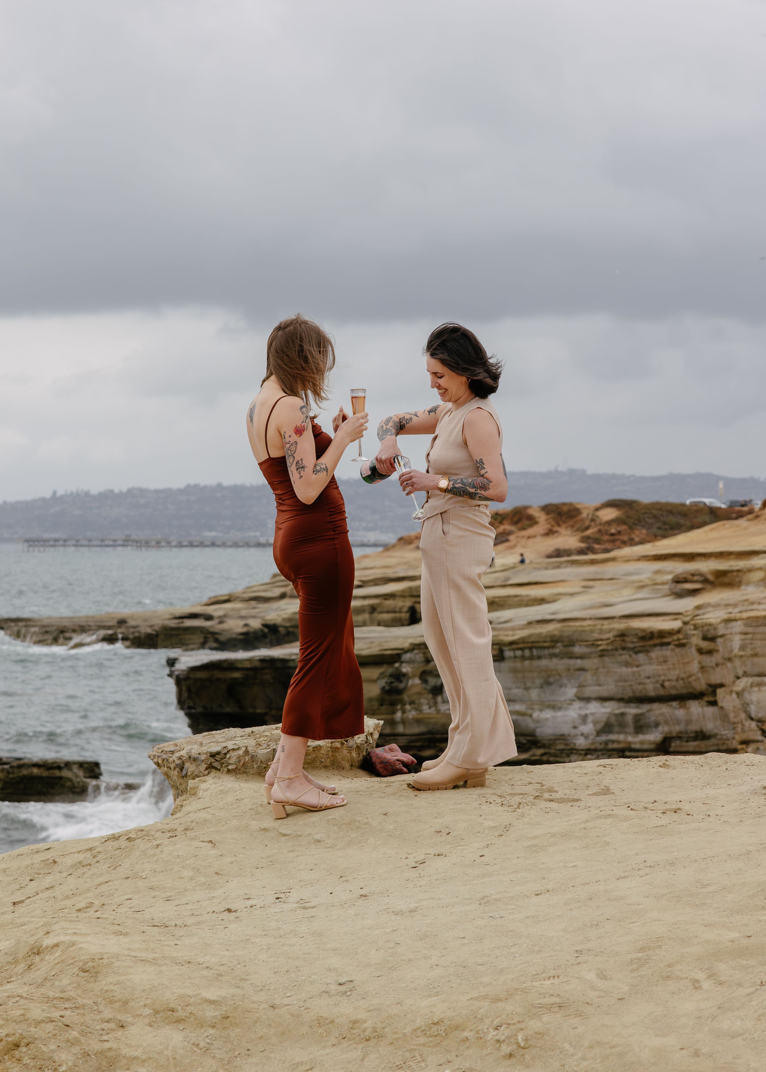 Two women standing on rocky coastal cliffs holding champagne glasses, one in red dress, one in beige pants.