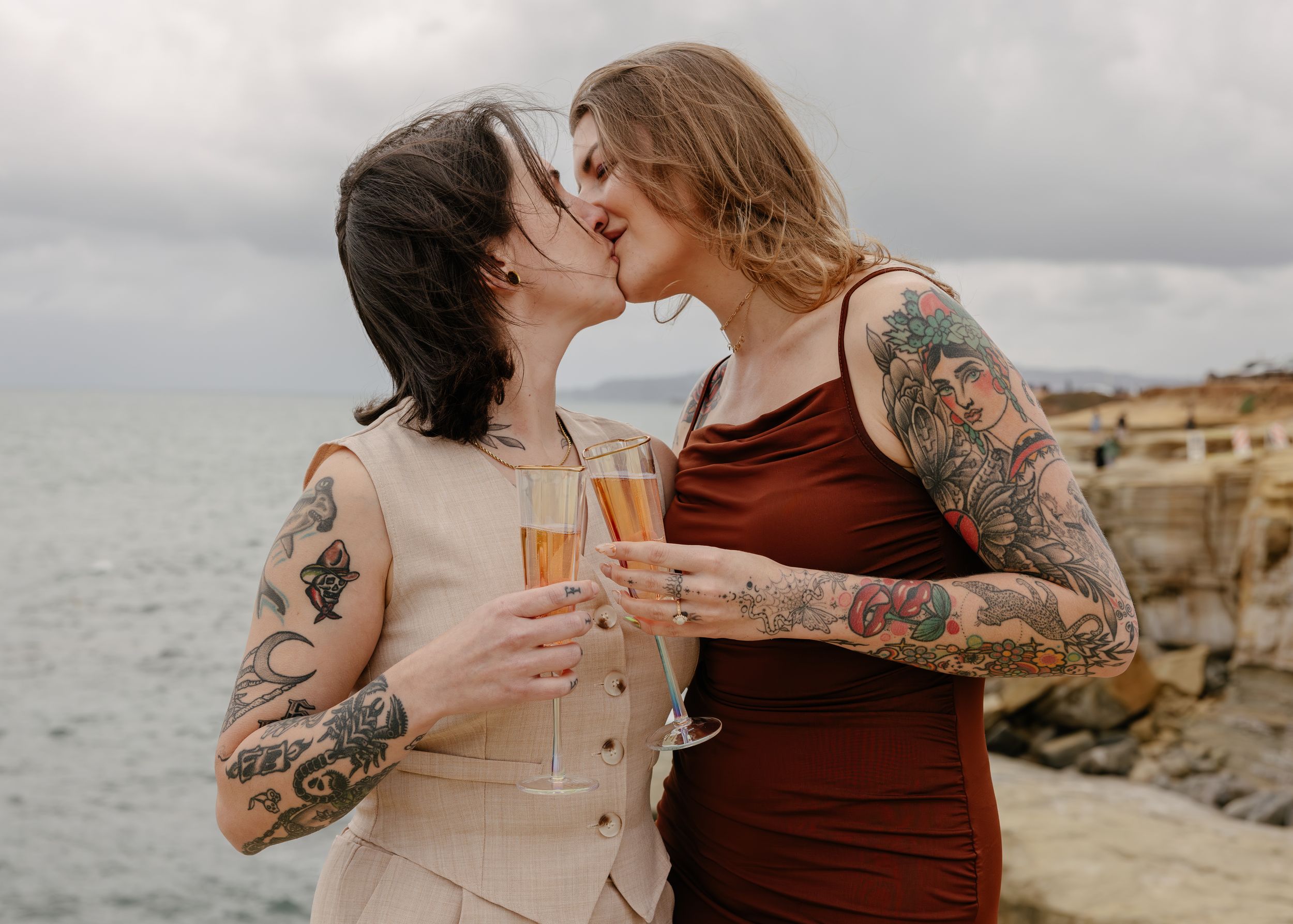 Two tattooed women sharing a kiss while holding champagne glasses outdoors by the ocean.
