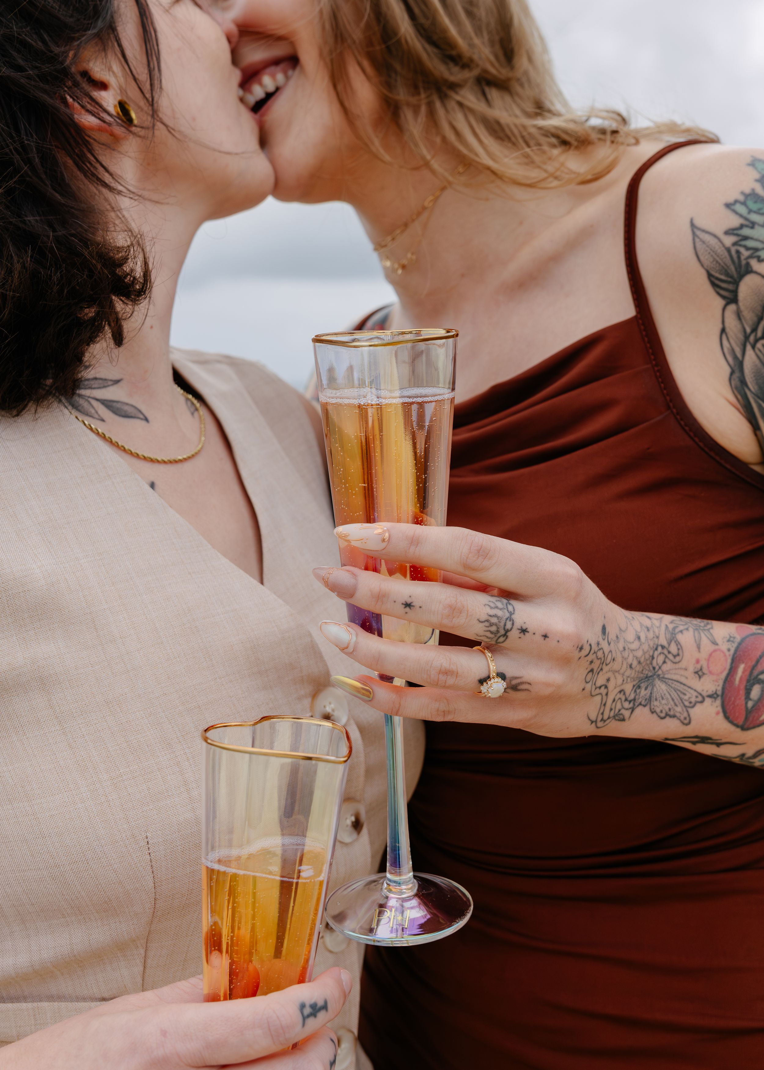 Two women with tattoos kissing and holding champagne flutes, celebrating a special occasion outdoors.