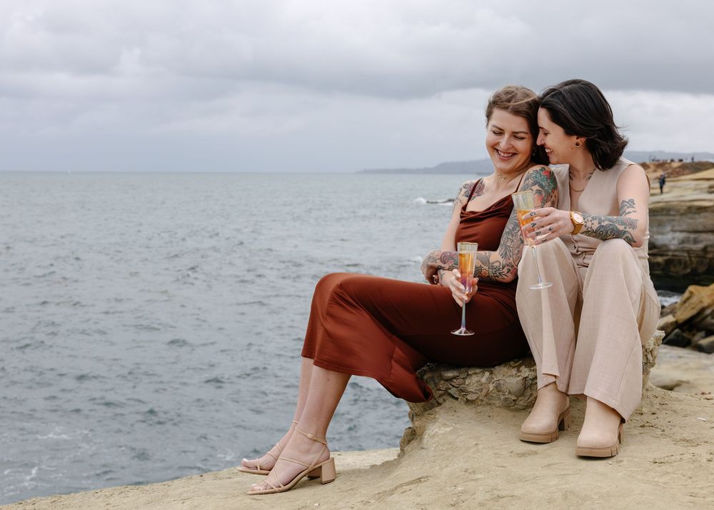 Two women sitting on coastal rocks, laughing and toasting with wine glasses by the ocean on a cloudy day.