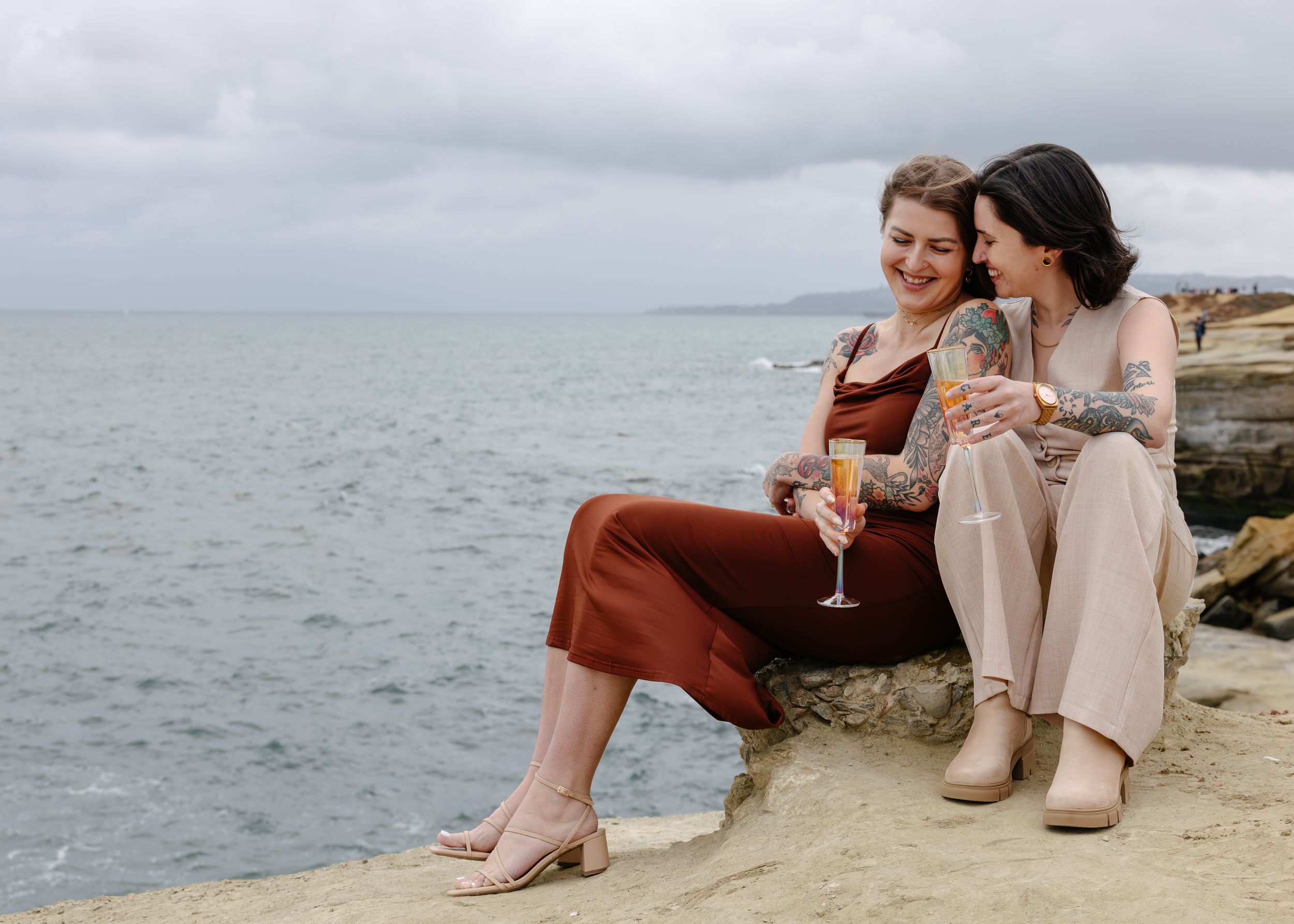 Two women sitting on coastal rocks, laughing and toasting with wine glasses by the ocean on a cloudy day.