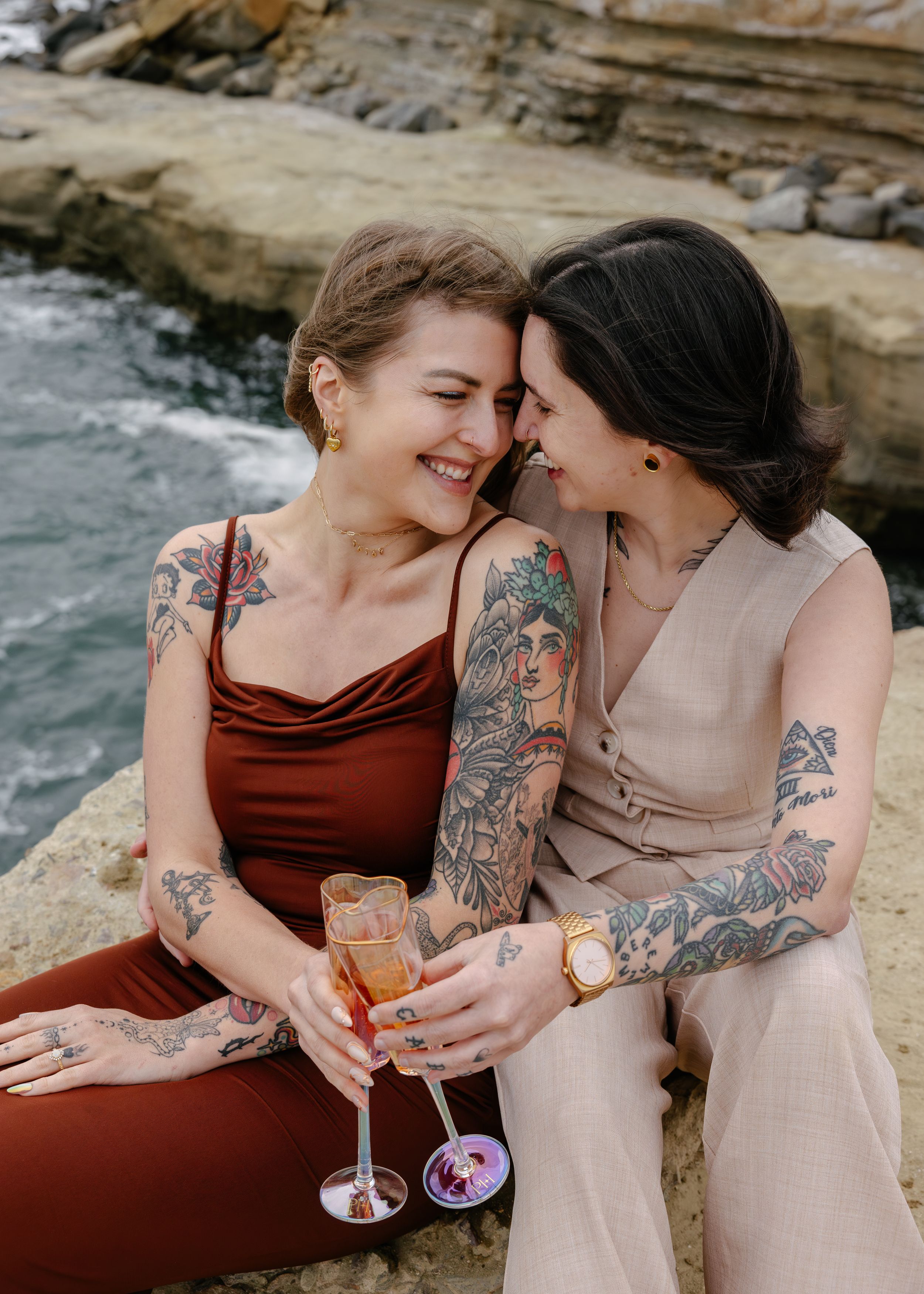 Two tattooed women in elegant dresses share a romantic moment on coastal rocks, holding champagne glasses.