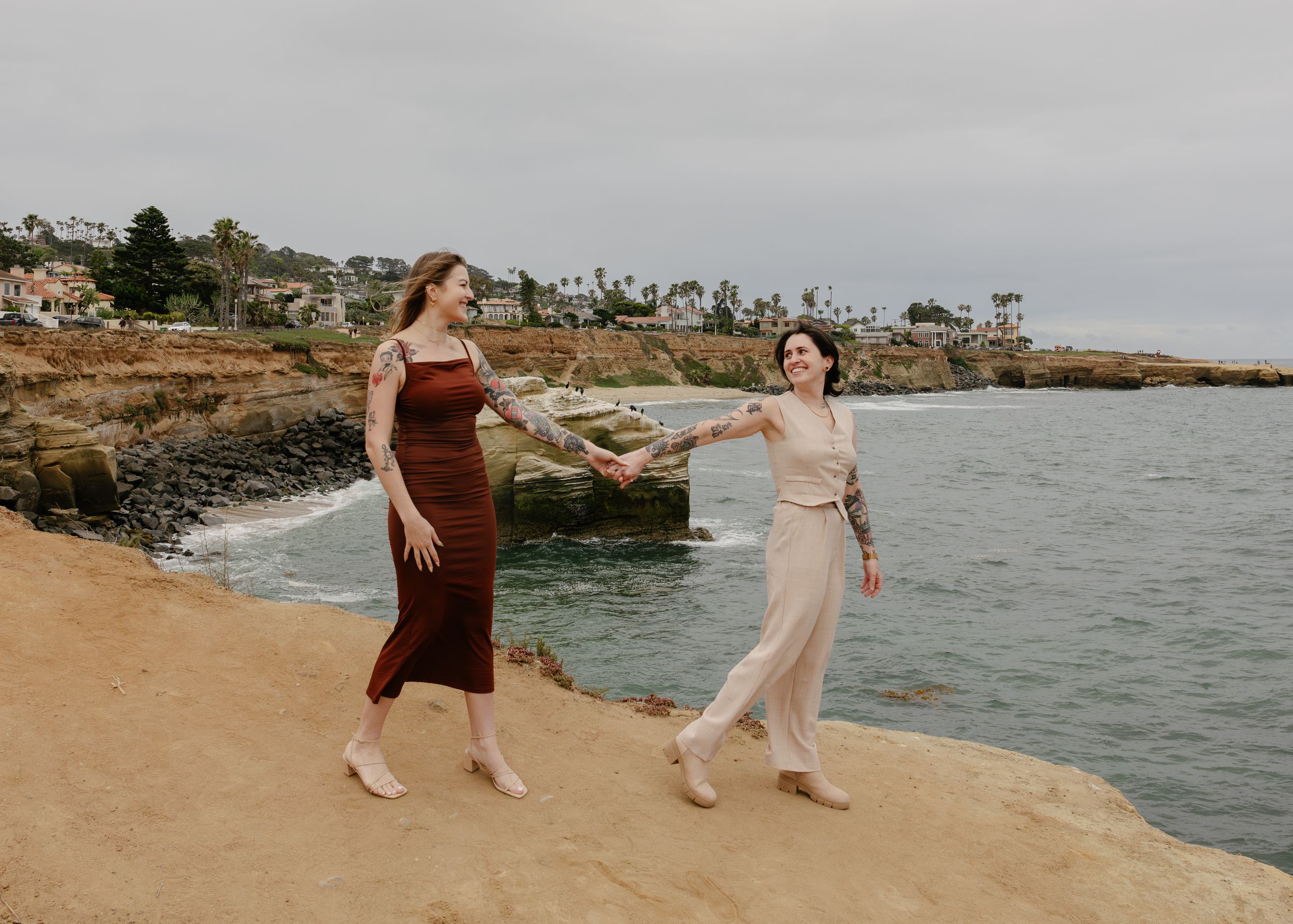 Two women in elegant dresses hold hands and dance on coastal cliffs overlooking the ocean in San Diego.