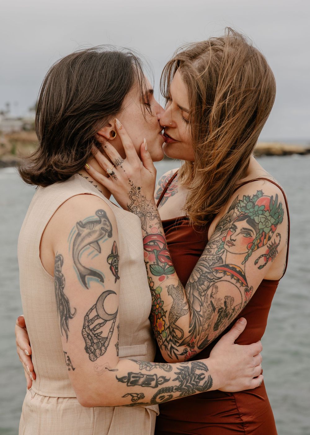 Two tattooed women sharing a romantic kiss outdoors near the ocean, embracing each other tenderly.