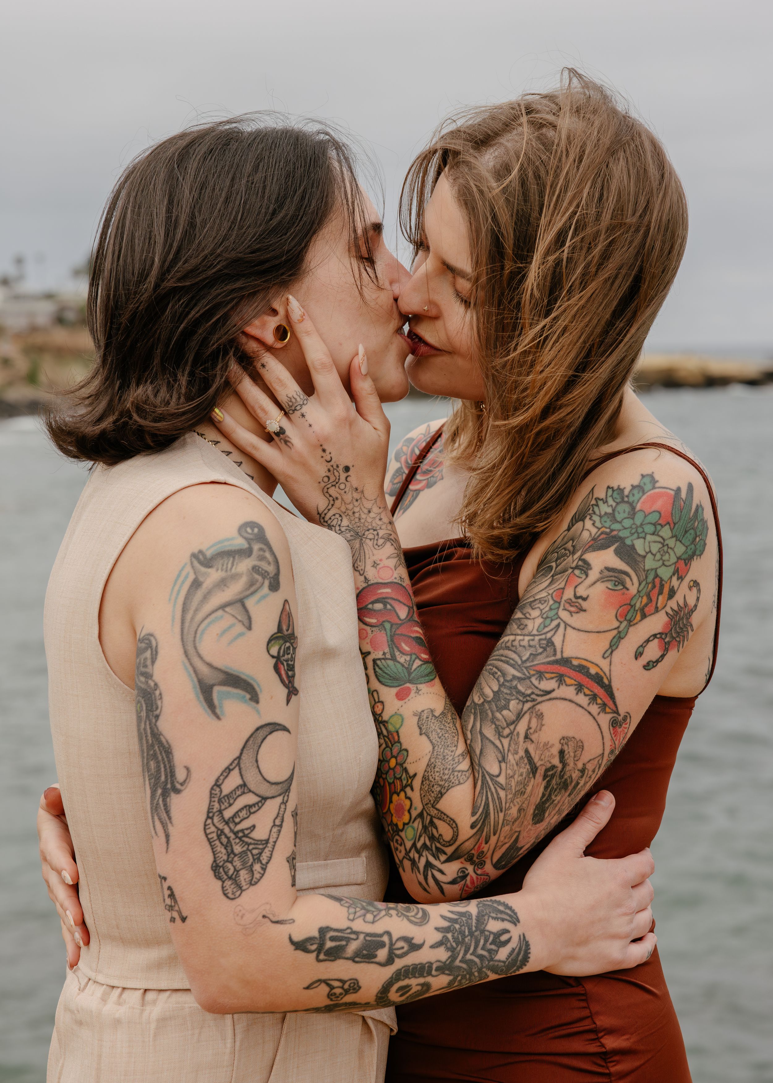 Two tattooed women sharing a romantic kiss outdoors near the ocean, embracing each other tenderly.