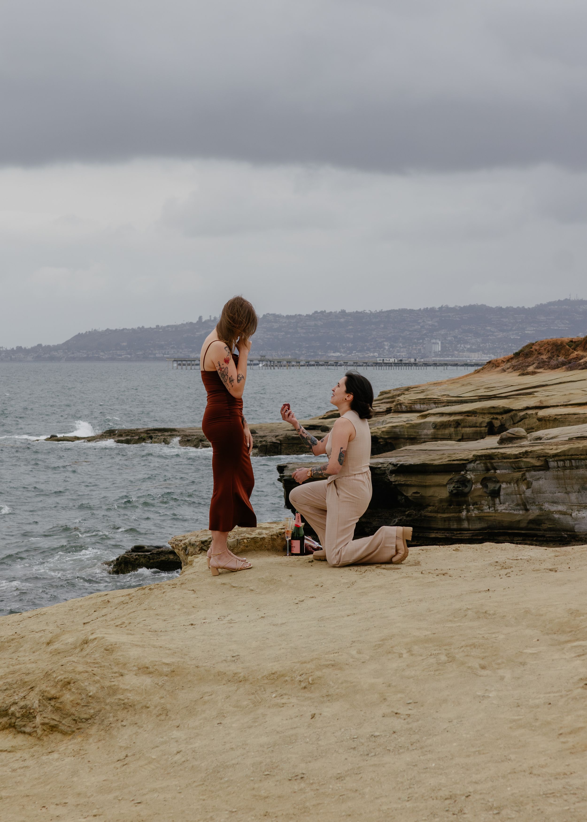 Person proposing on one knee to partner on rocky beach cliff overlooking ocean on cloudy day.