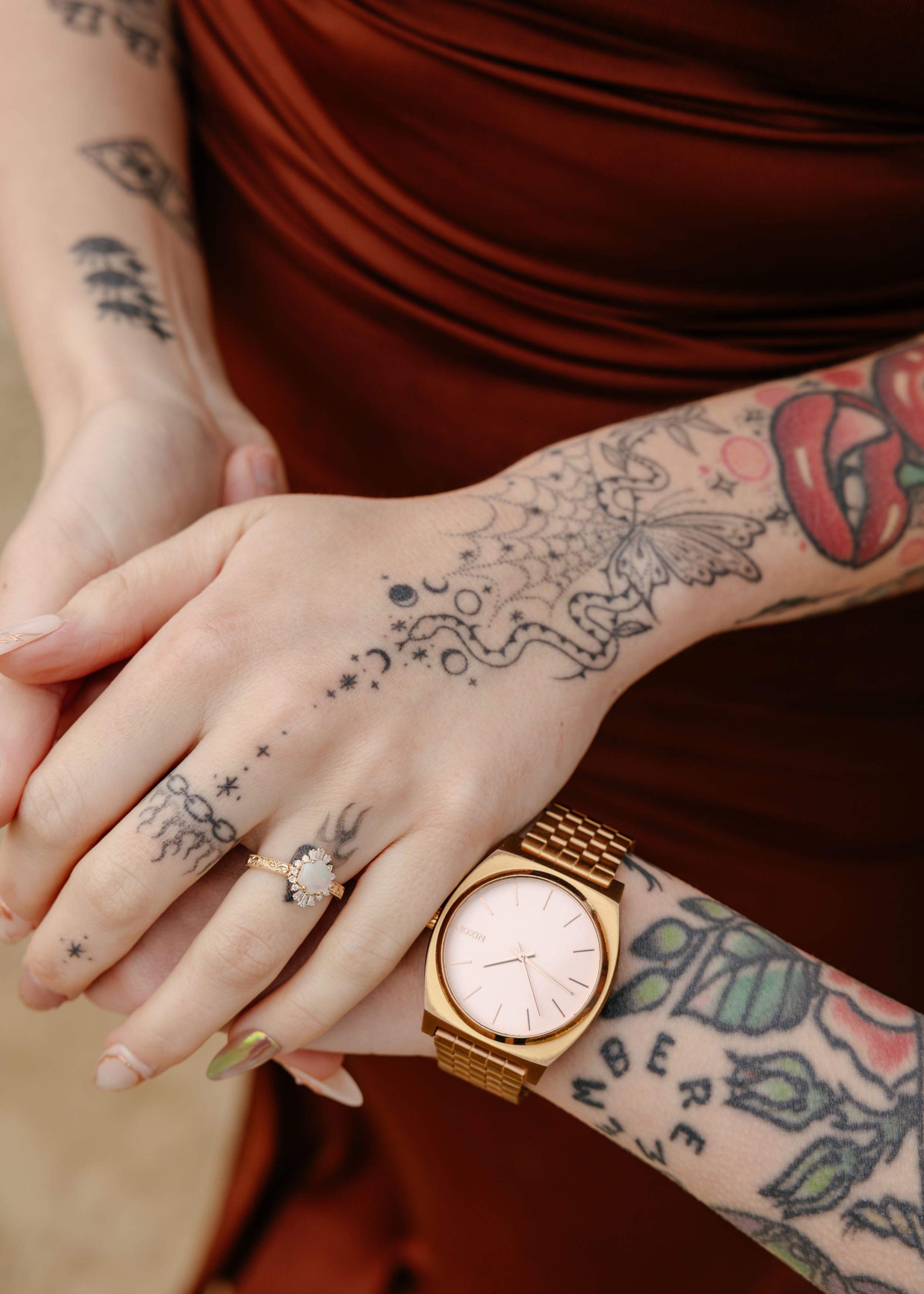 Tattooed woman wearing a gold Nixon watch and floral ring, hands clasped over a rust-colored satin dress.