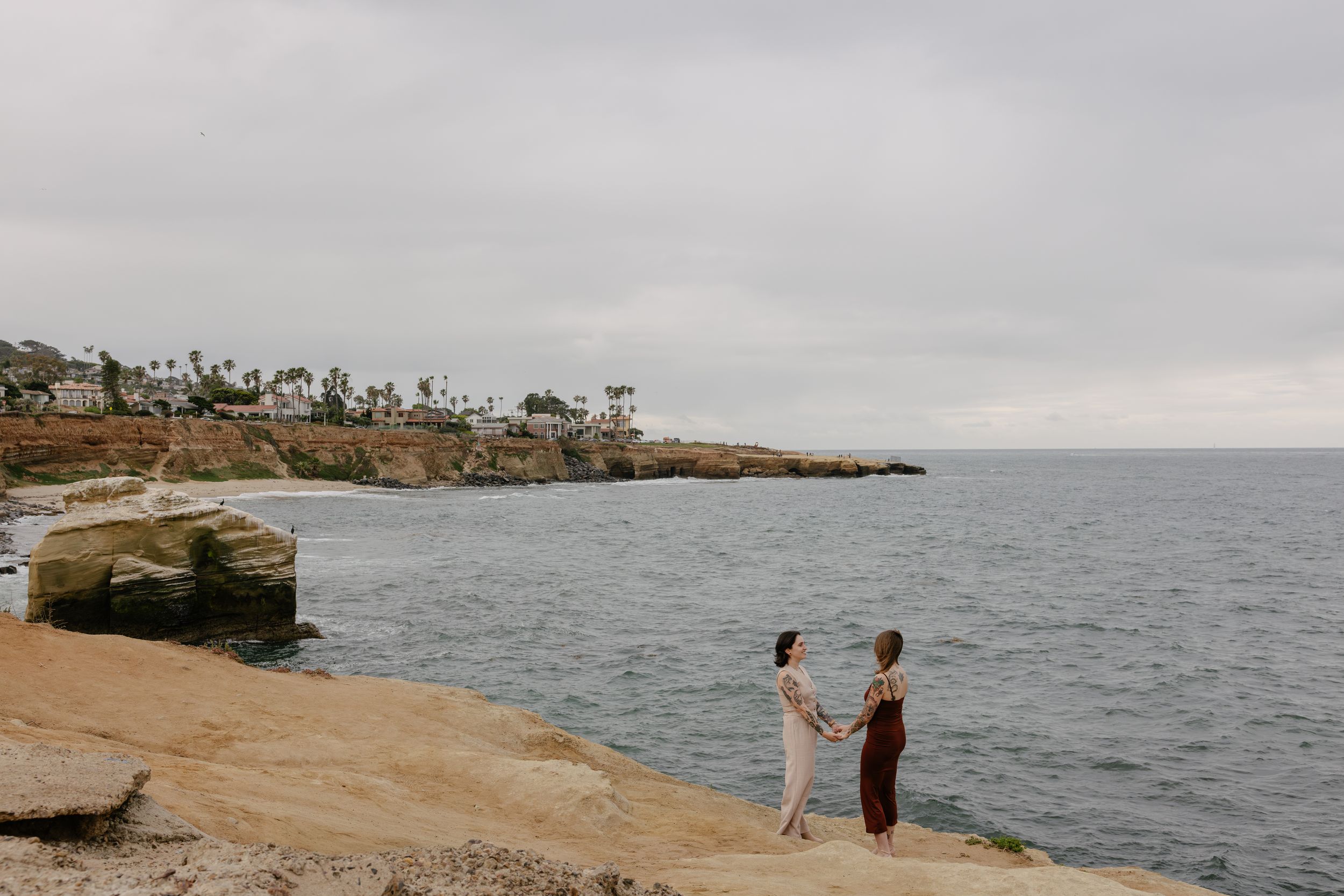 Two people hold hands on rocky coastal cliffs overlooking the ocean under an overcast sky.