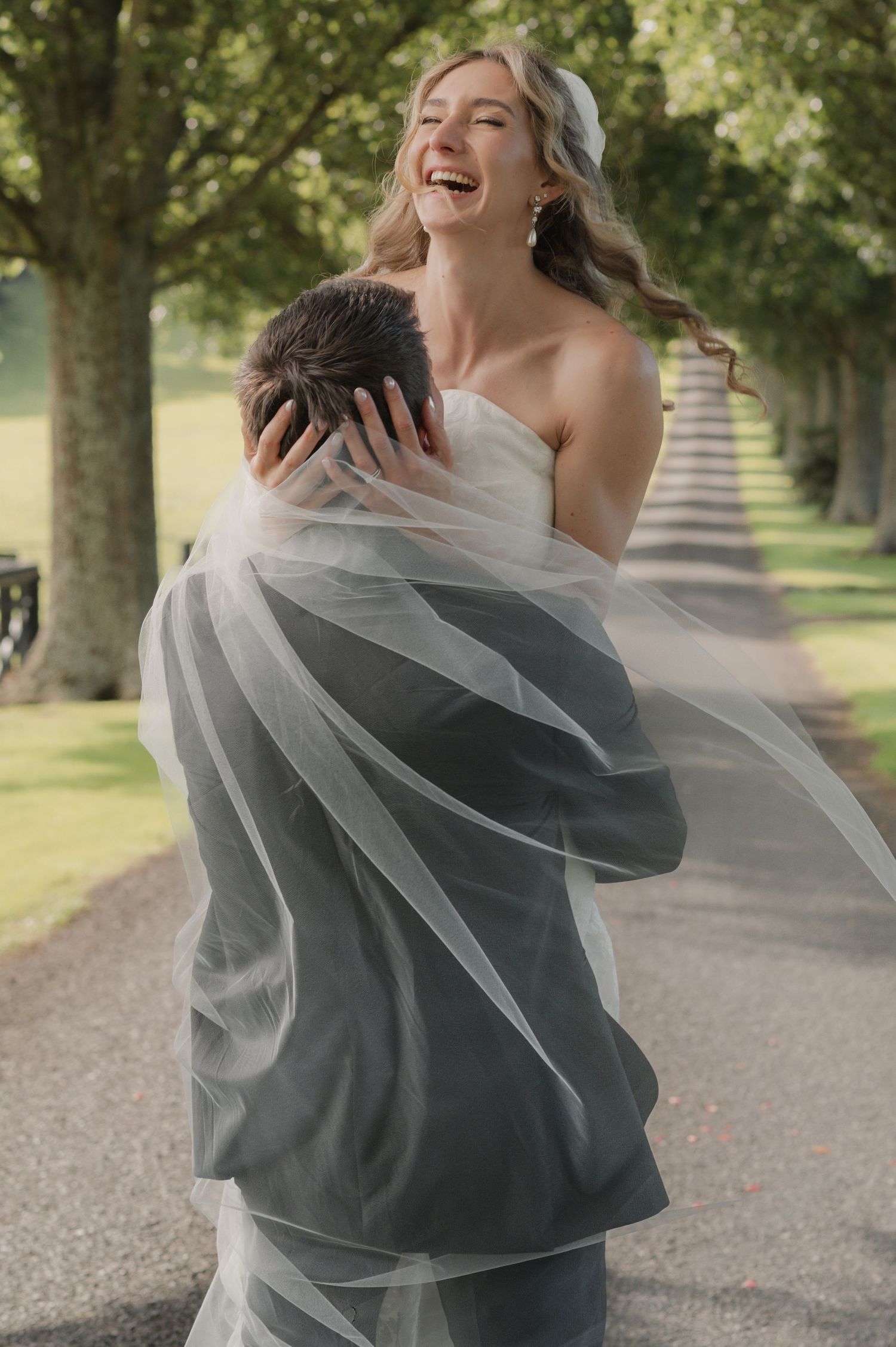 Joyful bride in white strapless gown and veil laughing while groom embraces her on a tree-lined path.