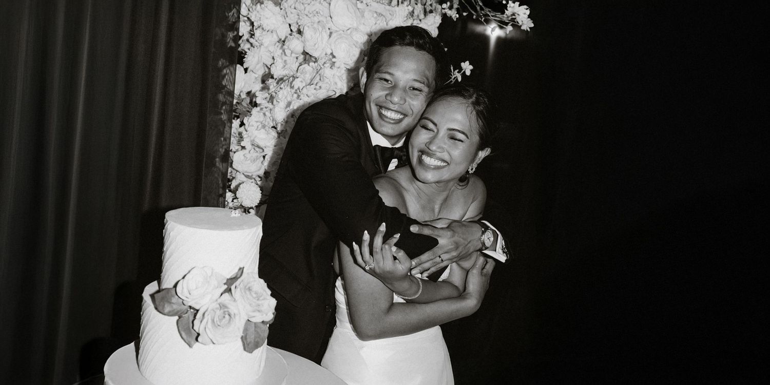 Bride and groom laughing beside their white wedding cake, black and white.