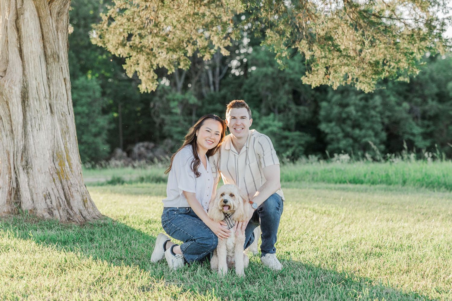 Couple smiling with golden doodle dog beside large tree in sunny green park outdoor portrait.