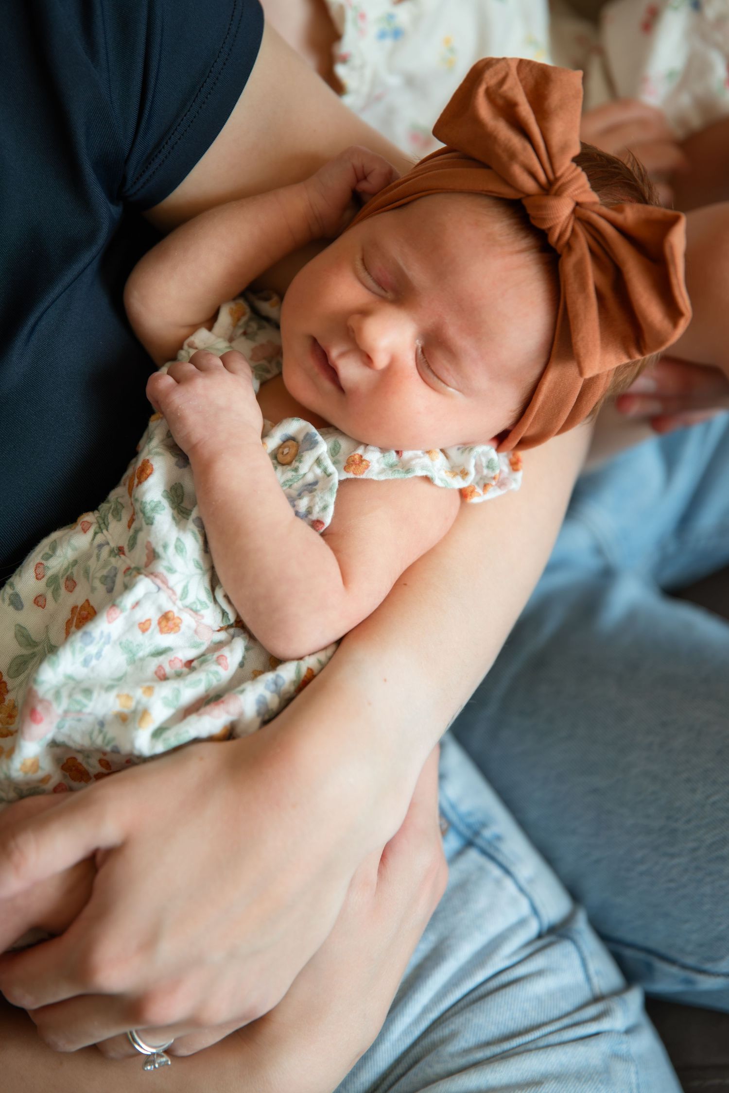 Newborn baby sleeping peacefully in parents arms wearing floral outfit and brown bow headband.