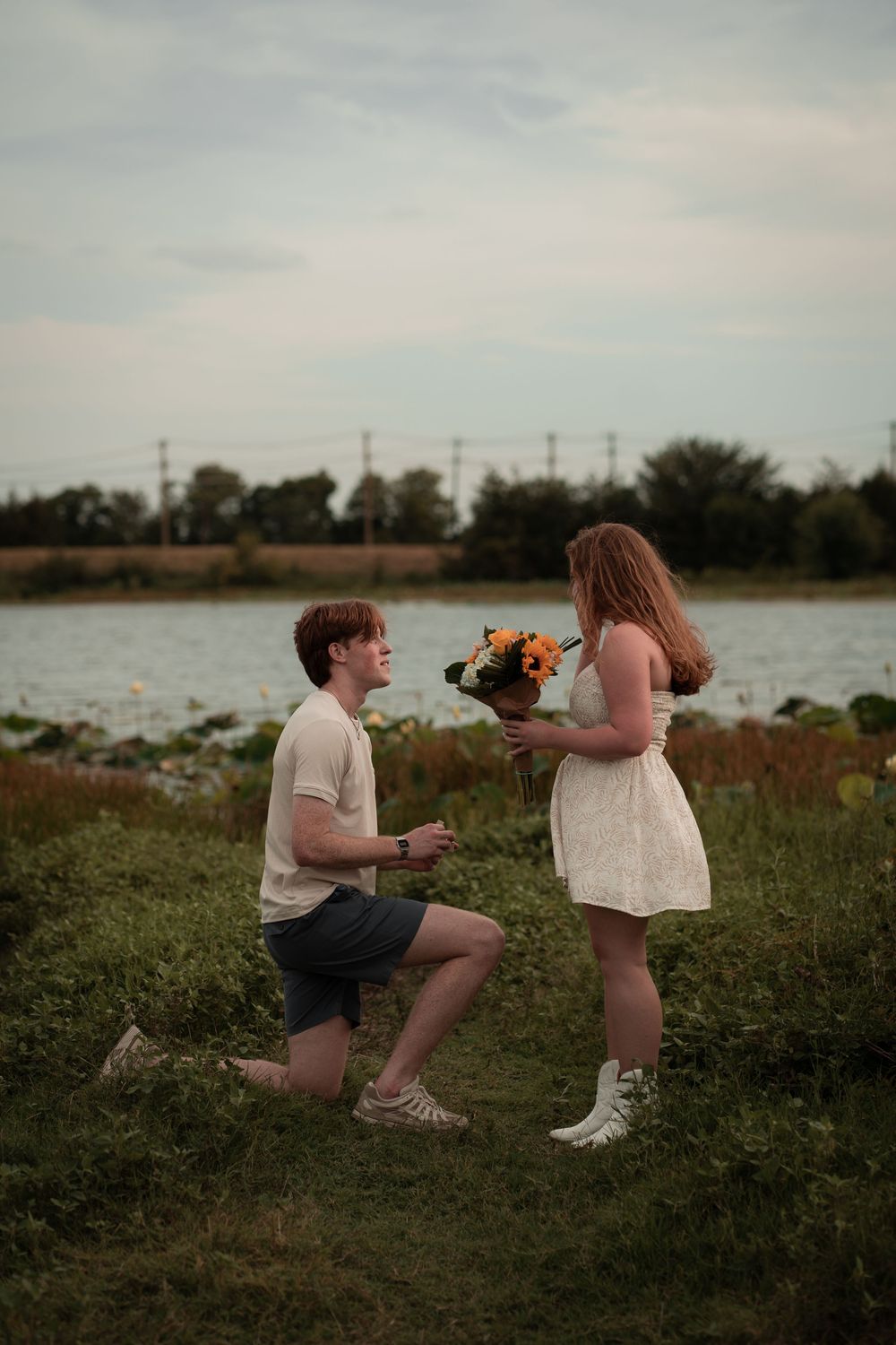 Young man kneeling on one knee presenting flowers to woman in white dress near a lake, romantic proposal scene outdoors.