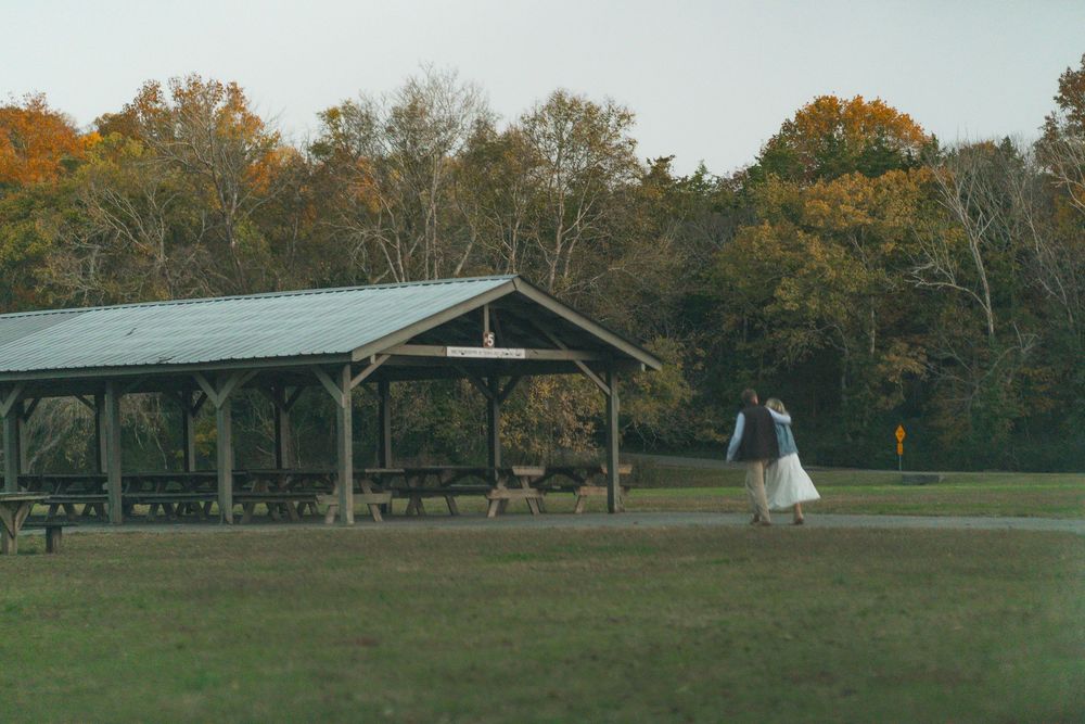 Person in white dress walking near a rustic open-air pavilion surrounded by autumn trees in a park.
