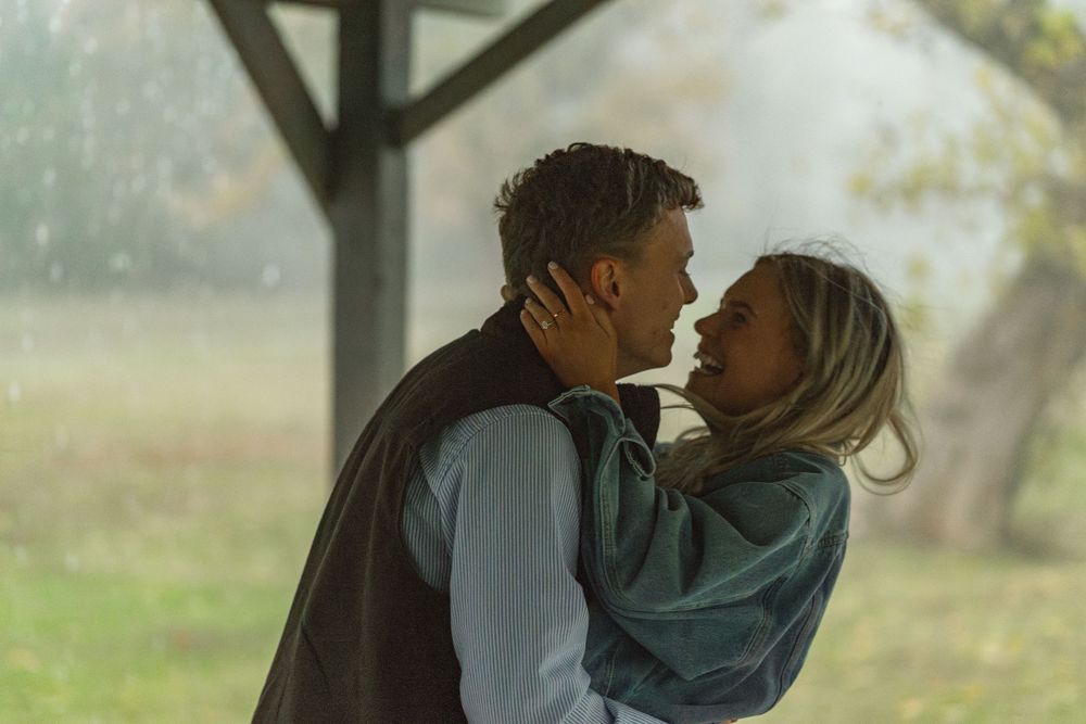 A couple shares a romantic moment in the rain, embracing and about to kiss outdoors near a tree.