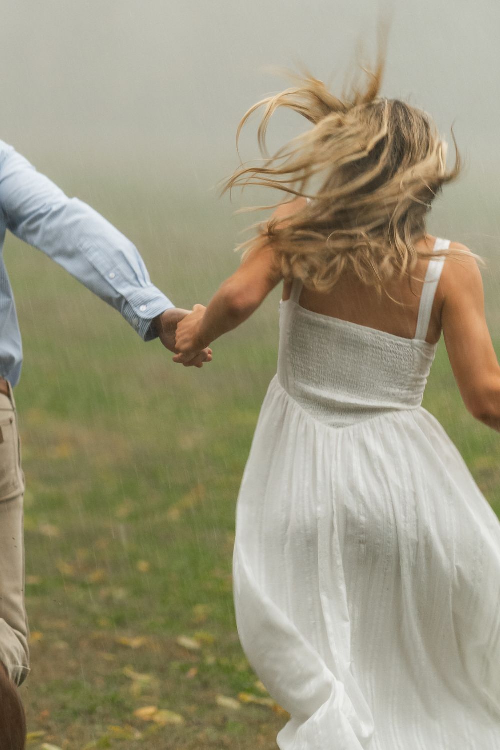 Couple holding hands walking through misty green field, woman in white dress with flowing blonde hair.