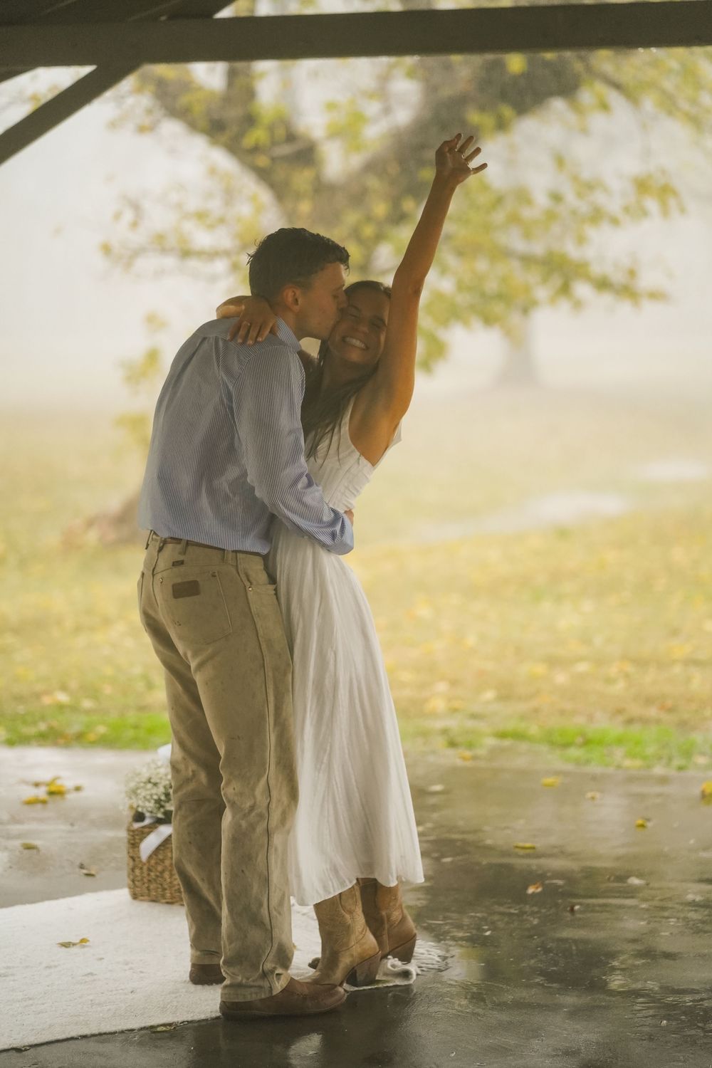 Couple dancing joyfully under a pavilion in misty park, woman in white dress with arm raised, man in blue shirt.
