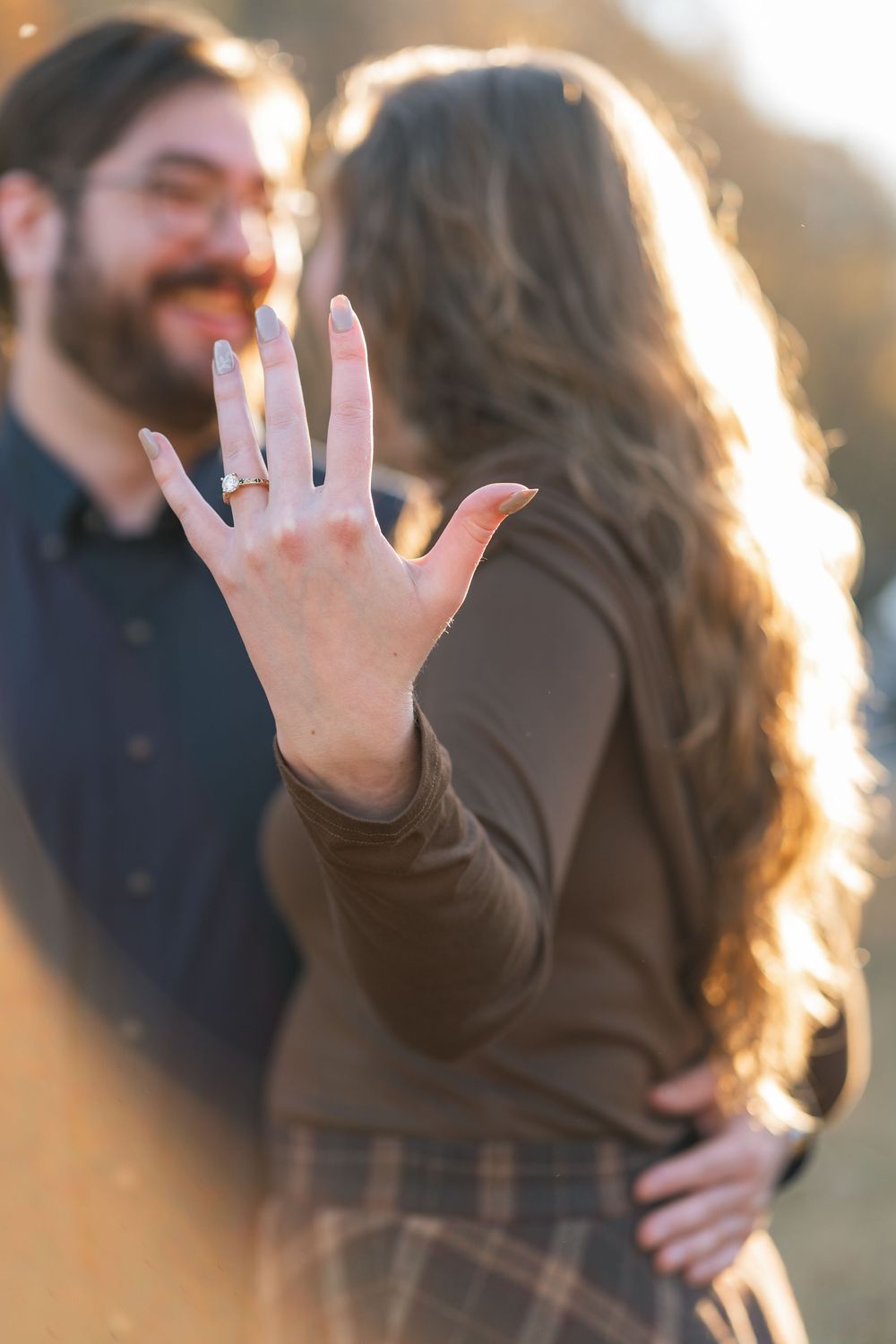 Couple showing off engagement ring outdoors in golden hour light, woman displaying diamond ring to camera.