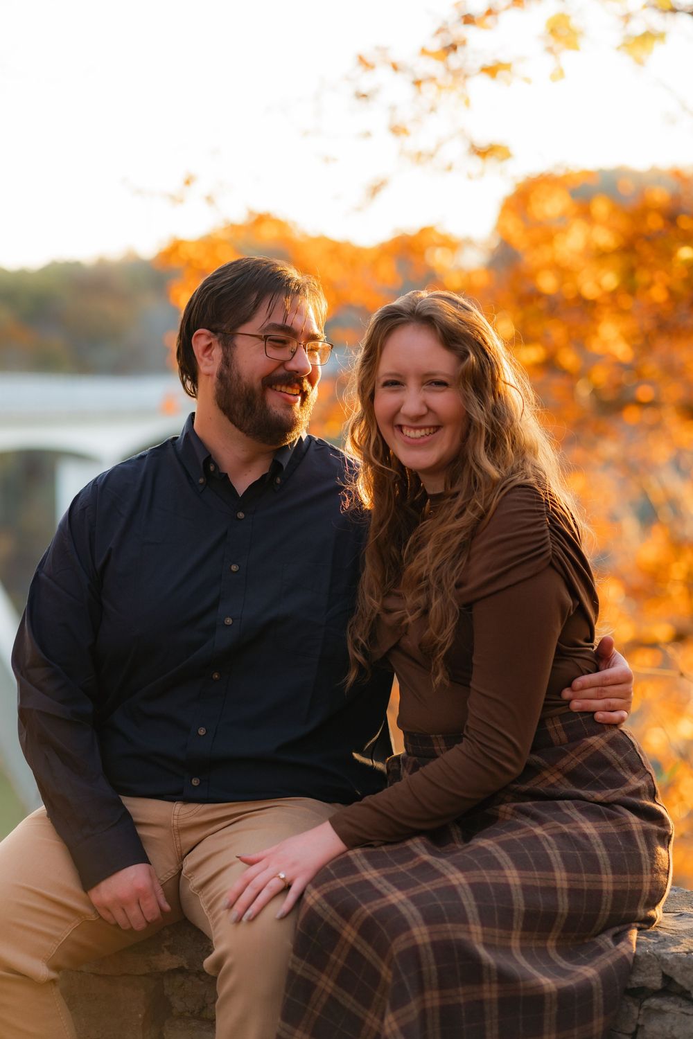 Couple smiling together outdoors during autumn, surrounded by vibrant orange foliage in a warm fall setting.