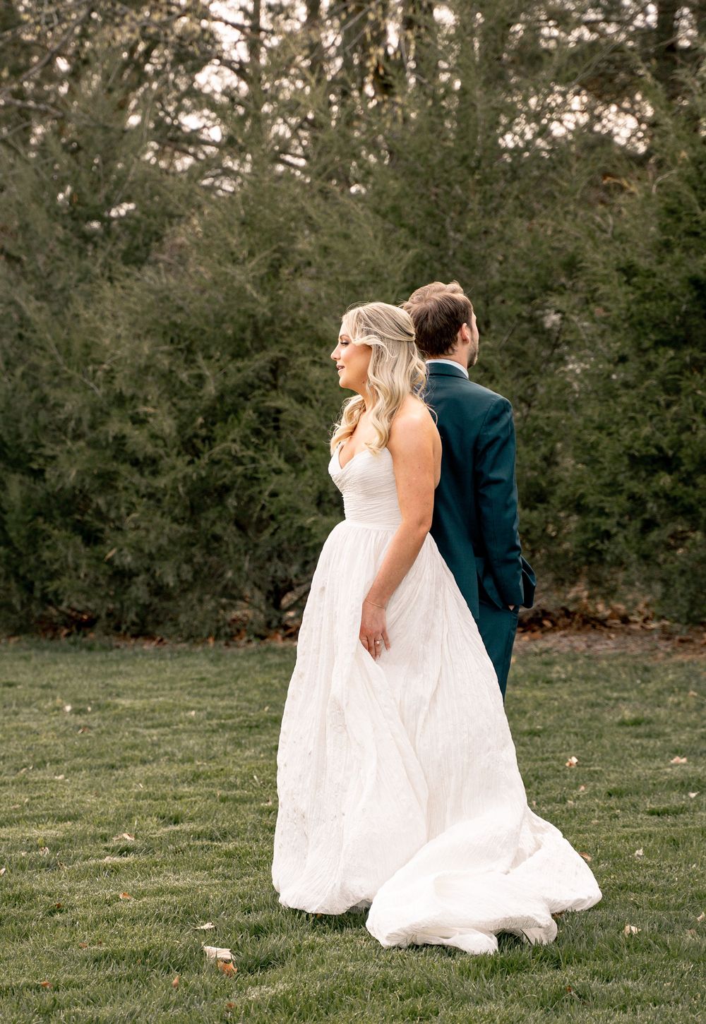 A bride in a white gown and groom in a dark suit walk together on a green lawn with tall evergreen trees behind them.