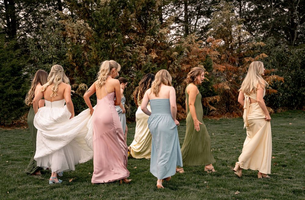 Six bridesmaids in pastel gowns walk across green lawn with backs to camera, surrounded by autumn trees.