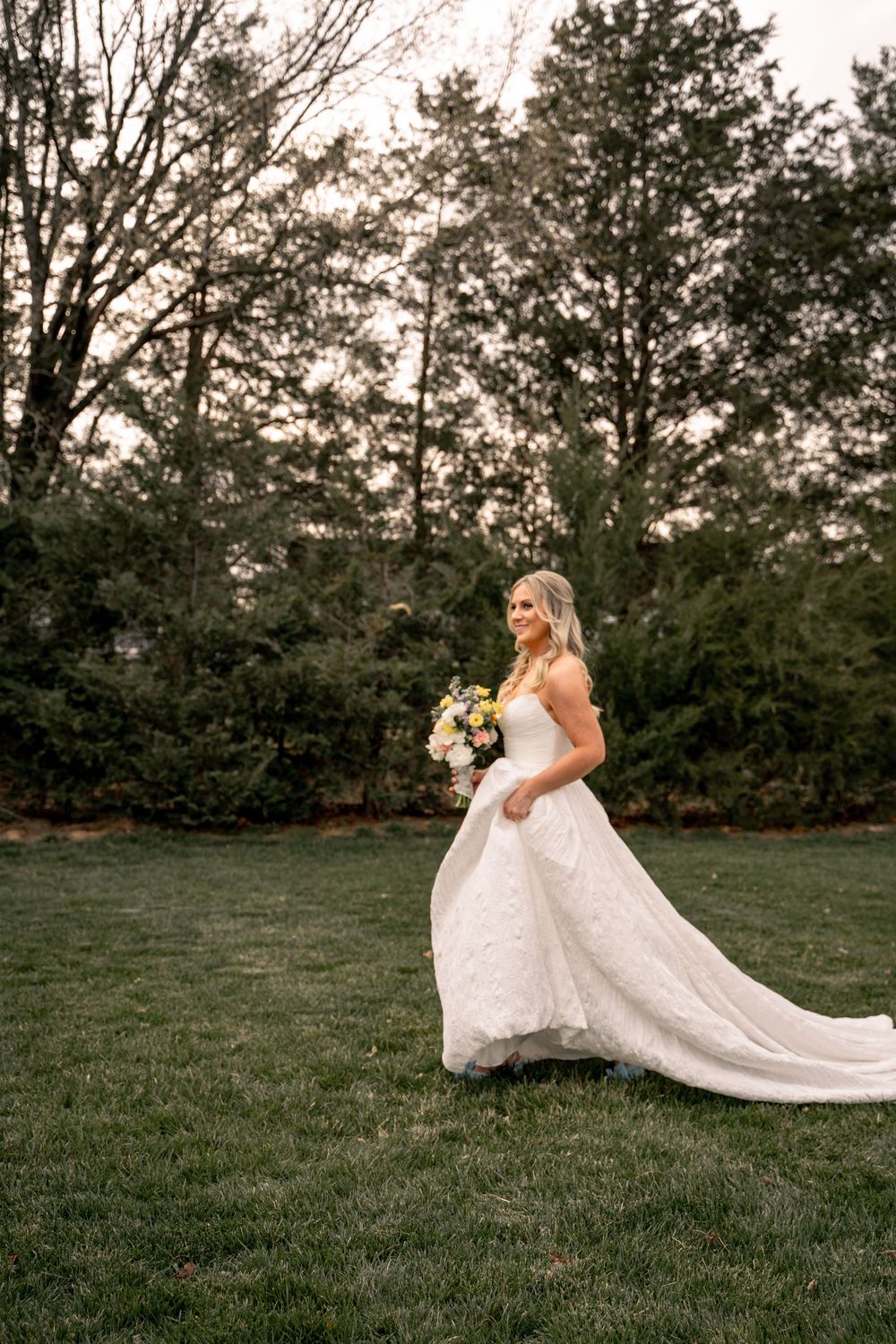 Bride in white lace ballgown holding colorful bouquet stands on green lawn with trees in background.