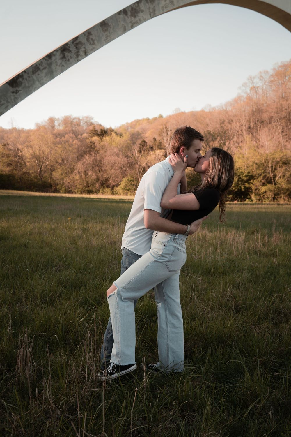 Couple sharing a romantic kiss in a green field with an arch and autumn trees at golden hour.