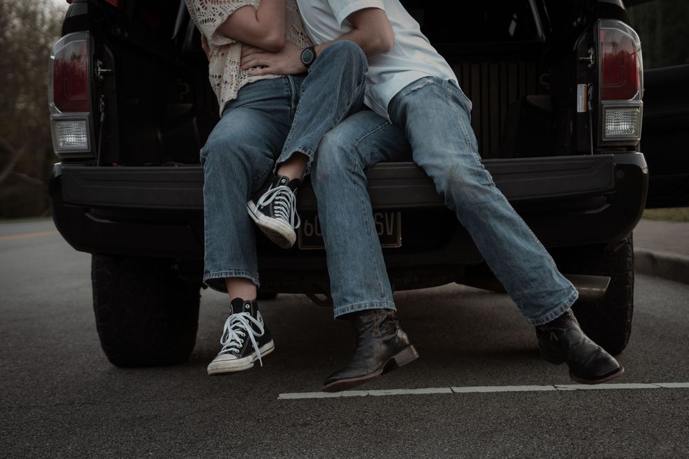 Two people in jeans sitting on a truck tailgate, one wearing Converse sneakers and the other in brown boots.