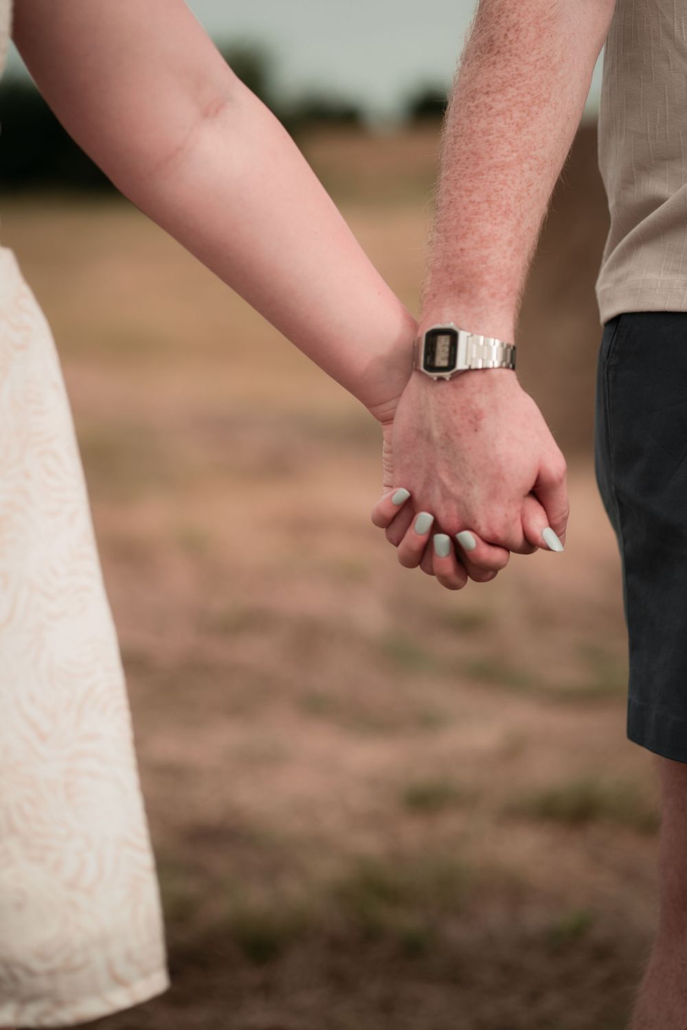 Couple holding hands outdoors in a field, woman with mint nails, man wearing a silver digital watch.