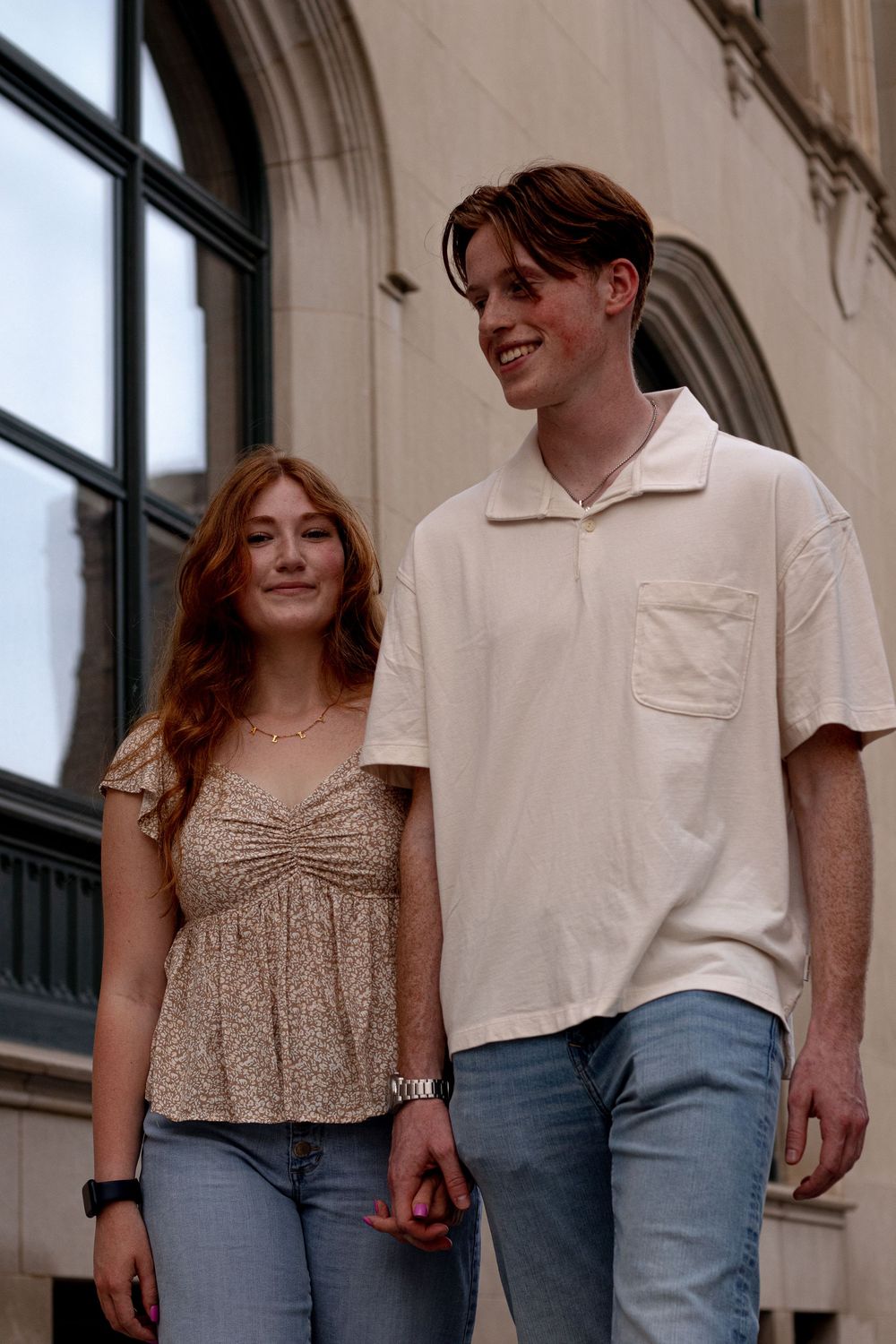 A young couple with red hair walks together outdoors, wearing casual summer outfits near a stone building.