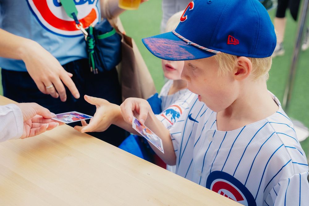 Young boy in Chicago Cubs jersey and cap examines baseball cards at a table during a fan event.