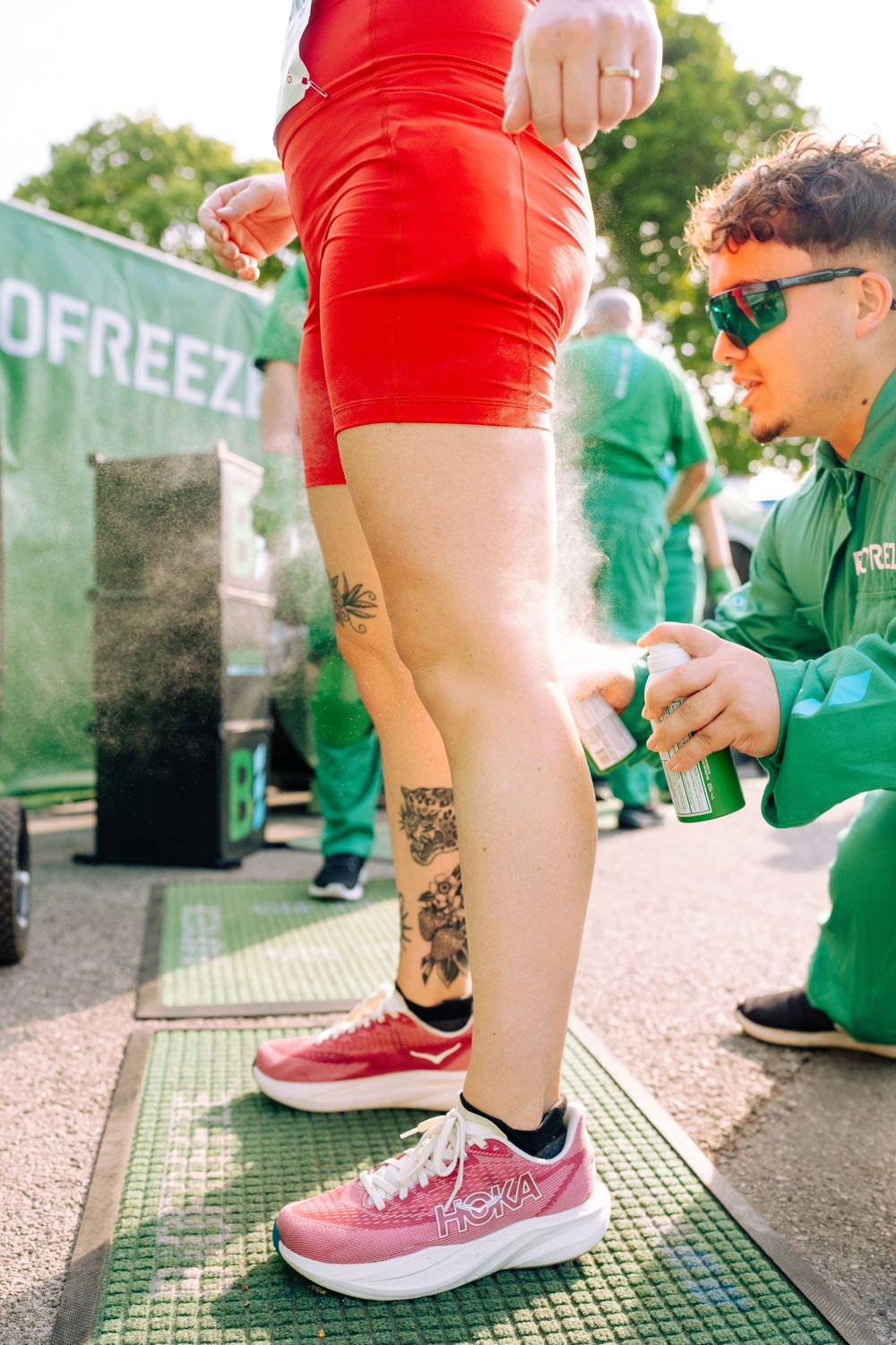 Person in red outfit with tattoos getting legs sprayed with freeze spray by worker in green uniform at outdoor event.