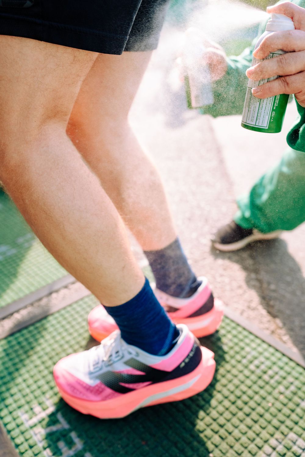 Runner wearing bright pink sneakers and blue socks gets spray applied to legs before a race.