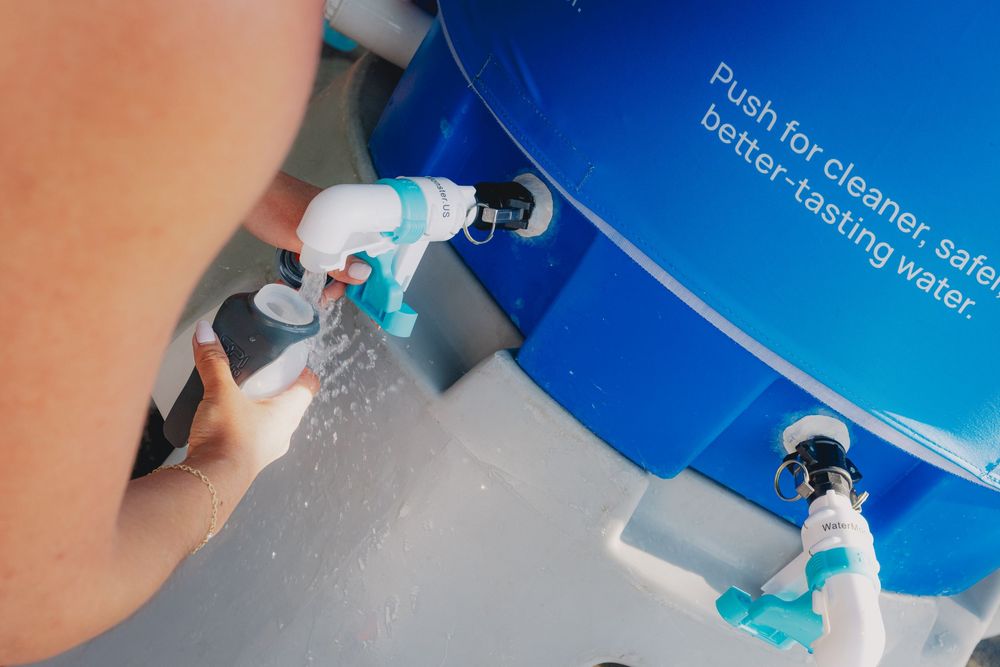 Person filling water bottle at blue filtered water station with push-button dispenser for clean, safe drinking water.