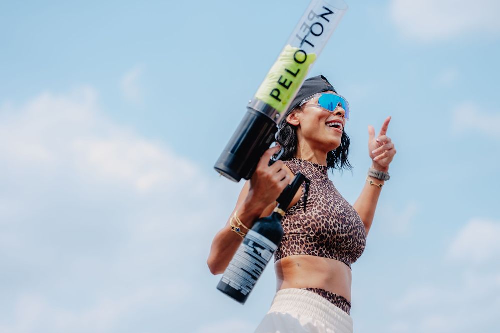 Woman in leopard print top holds Peloton cannon, giving thumbs up outdoors under blue sky.